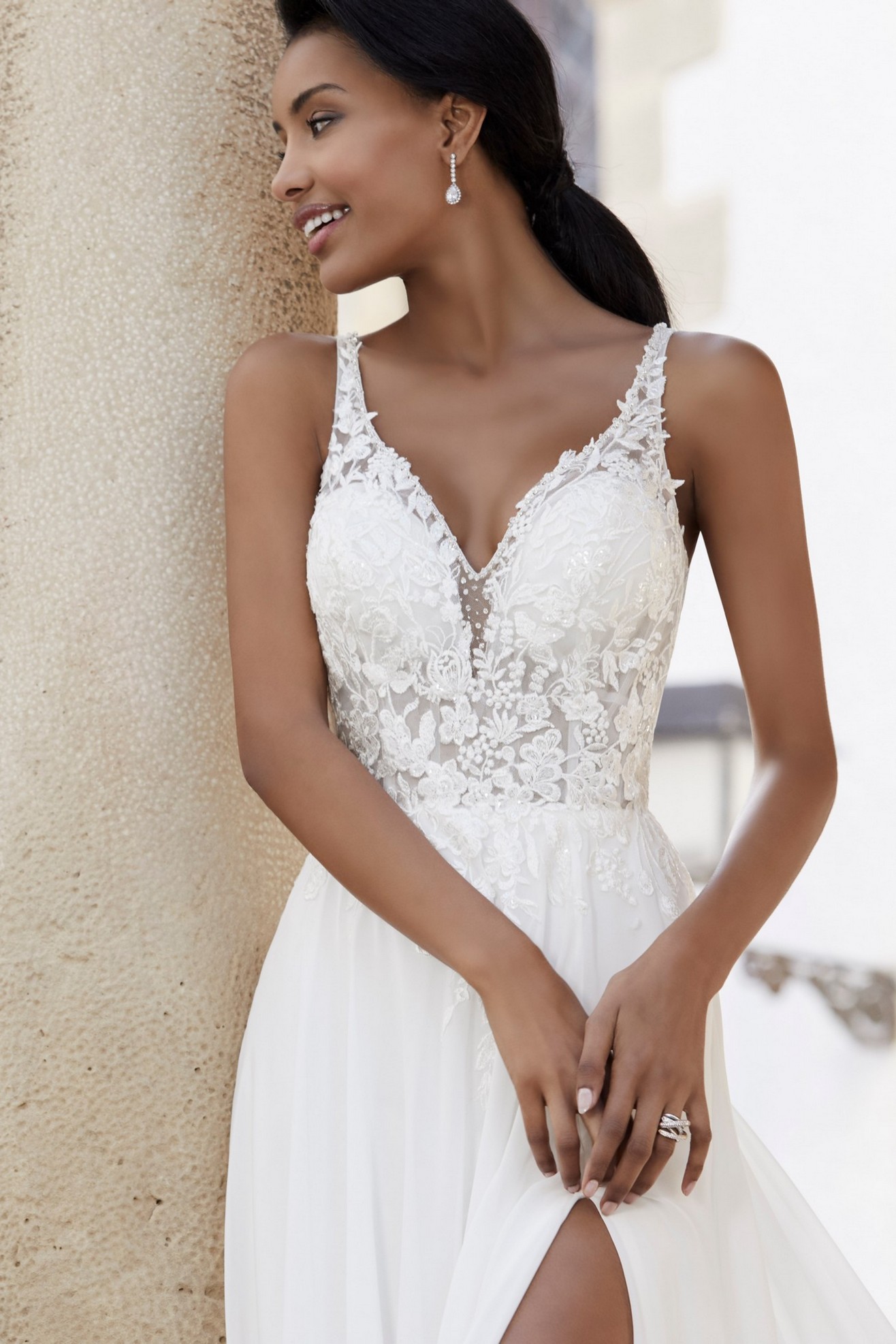 Woman standing smiling next to pillar wearing lace wedding dress with v-neckline and thigh split detail